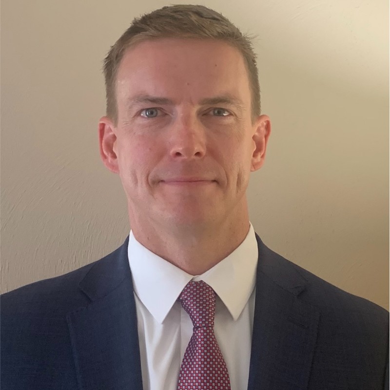 A professional headshot of a man with short light brown hair, wearing a dark suit, white dress shirt, and a red patterned tie. He is looking directly at the camera with a neutral expression and is standing against a plain light-colored background.