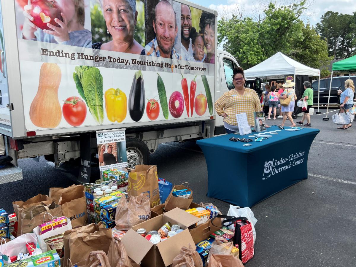 A woman stands next to a booth with a table covered in pamphlets and promotional items. Behind her is a truck with a colorful banner displaying vegetables and a message about ending hunger.