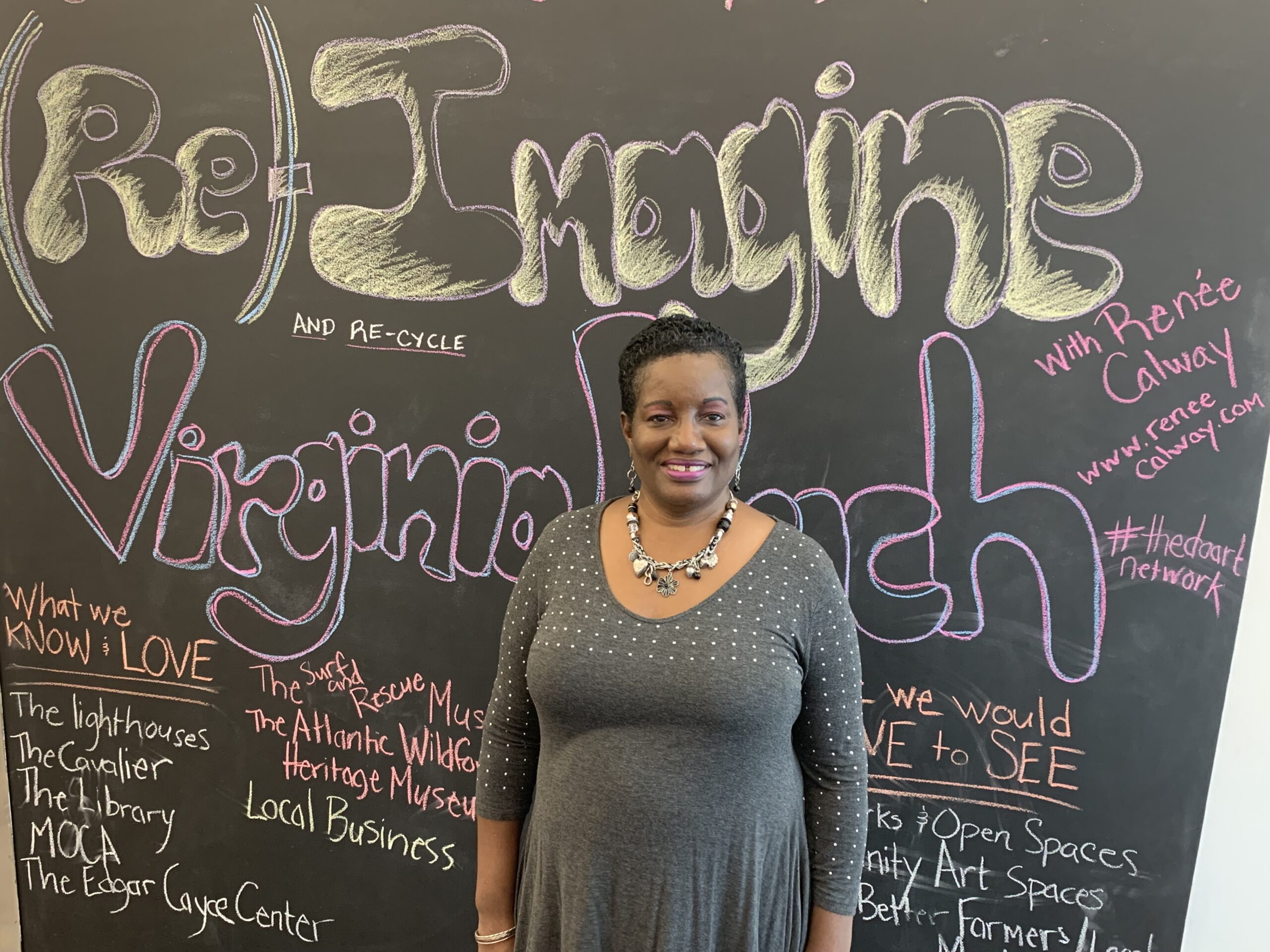 A woman stands smiling in front of a blackboard covered with colorful chalk writing. The board reads 'Re-Imagine Virginia Beach' in large letters, with other smaller phrases and notes around it. The woman is wearing a gray dress with silver polka dots and a statement necklace.